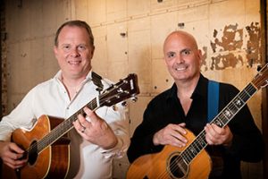 Two men standing next to each other, both holding guitars