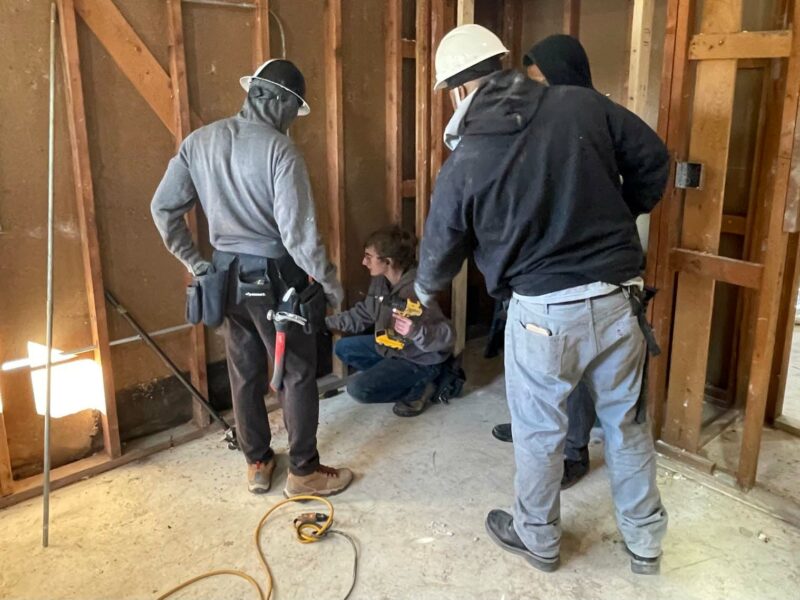 Three workers standing around a kneeling fourth person looking at an open wall.