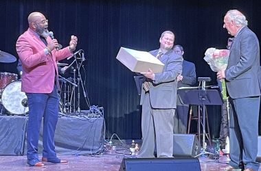 man holding flowers being presented with congratulatory cake by two other men, one holding a microphone, the other holding the cake