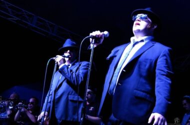 two men wearing suits, hats and sunglasses singing behind microphones